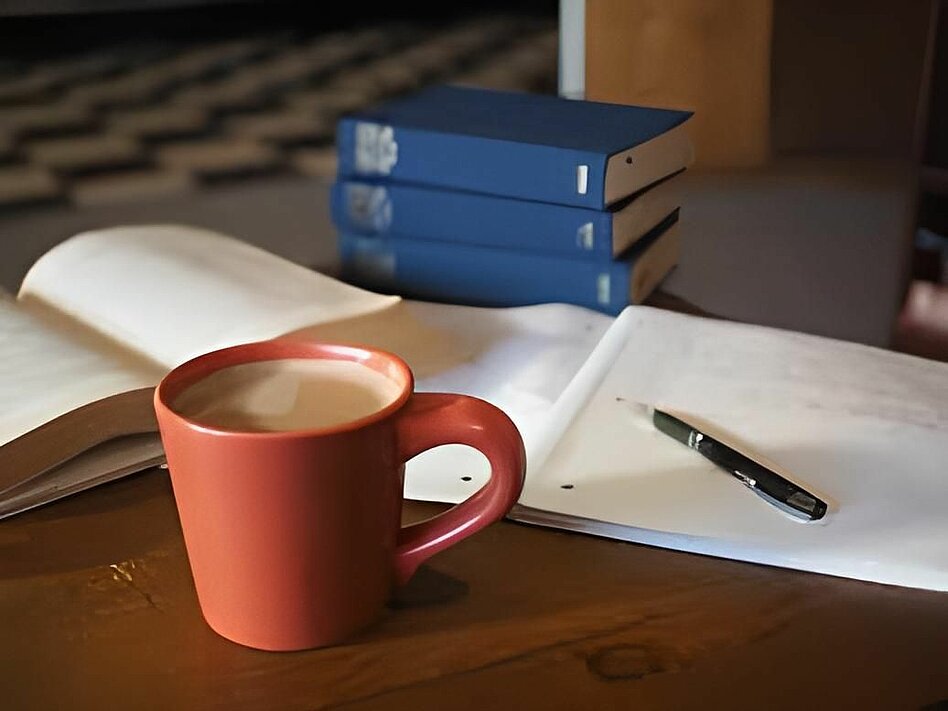 bureau de travail avec des livres, un stylo posé sur un cahier et une tasse de café au premier plan.